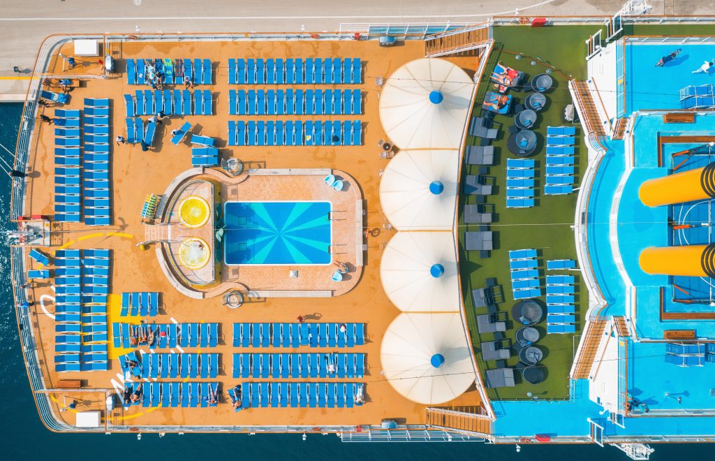 Aerial view of swimming pool, people on sunbeds, umbrellas on the cruise ship in summer. View from above of relaxing people on deck chairs, blue water in pool, wooden deck. Luxury cruise liner. Resort
